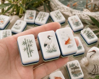 May include: A set of rectangular mahjong tiles featuring white surfaces with detailed illustrations of bamboo stalks, a crane, and goldfish. Each tile has a layered base with blue and green accents, showcasing intricate nature-themed artwork.