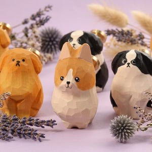 May include: A collection of hand-carved wooden dog figurines in shades of tan, black, and white. The dogs are displayed on a lilac surface, accompanied by dried lavender and decorative elements.