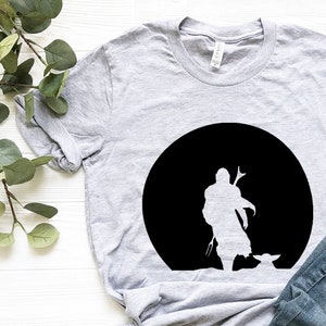 May include: A light gray t-shirt featuring a black silhouette of a character and a small creature against a black circle. The shirt is paired with blue jeans and brown sandals with black straps. The shirt is made of a soft material.