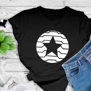 Puede incluir: Camiseta negra con un dise&ntilde;o de estrella blanca dentro de un c&iacute;rculo. La imagen tambi&eacute;n incluye pantalones cortos de mezclilla, gafas de sol, un tel&eacute;fono inteligente y zapatillas deportivas, con una peque&ntilde;a planta en una superficie de madera.