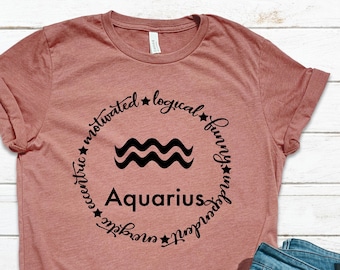 以下が含まれることがあります： 「Aquarius」の文字と黒い波のシンボルが入ったダスティローズのTシャツ。シンボルは「やる気、論理的、面白い、自立、エネルギッシュ、風変わり」という言葉と星で囲まれています。水色のスニーカーとブルージーンズも画像にあります。
