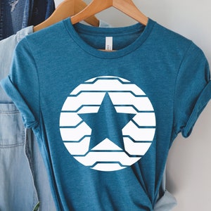 Puede incluir: Camiseta de color turquesa con un dise&ntilde;o de estrella blanca dentro de un c&iacute;rculo. La estrella es s&oacute;lida y el c&iacute;rculo est&aacute; formado por l&iacute;neas horizontales. La camiseta est&aacute; colgada en una percha de madera.