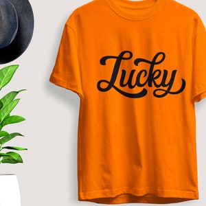 May include: Bright orange t-shirt with the word "Lucky" in black script. The shirt is displayed on a white background, with a dark blue hat and a green plant in a white pot in the background.