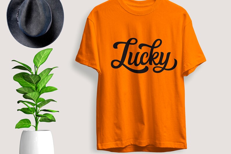 May include: Bright orange t-shirt with the word "Lucky" in black script. The shirt is displayed on a white background, with a dark blue hat and a green plant in a white pot in the background.