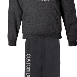 Pode incluir: Conjunto de moletom com capuz e cal&ccedil;as de moletom cinza carv&atilde;o. O moletom com capuz apresenta o texto "CUSTOM DESIGN" no peito, e as cal&ccedil;as de moletom t&ecirc;m o mesmo texto verticalmente na perna esquerda. Uma roupa confort&aacute;vel e casual.
