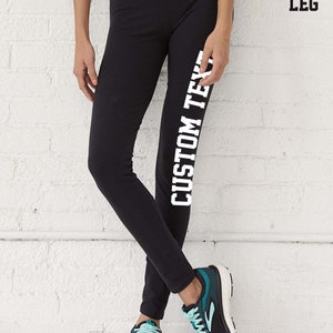 May include: Black leggings with the text "CUSTOM TEXT" printed vertically in white on the left leg. The person is wearing black and teal athletic shoes. The background is a white brick wall.