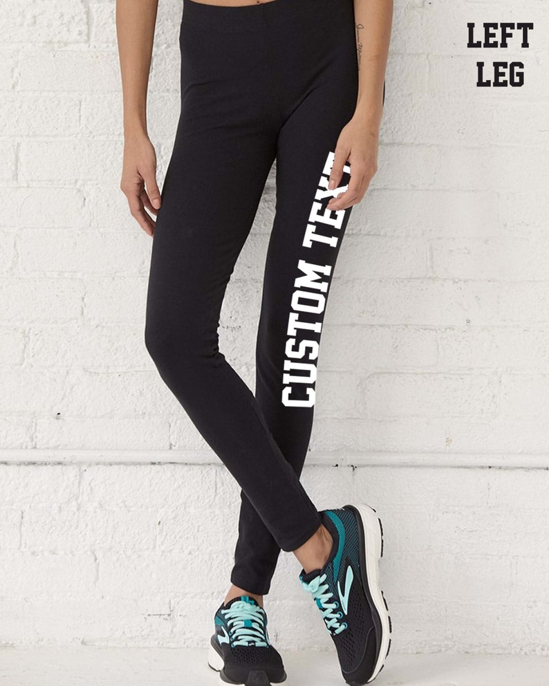 May include: Black leggings with the text "CUSTOM TEXT" printed vertically in white on the left leg. The person is wearing black and teal athletic shoes. The background is a white brick wall.