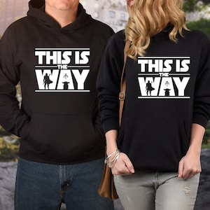 Puede incluir: Sudaderas con capucha negras con la frase "THIS IS THE WAY" en letras blancas. El dise&ntilde;o incluye una silueta de personaje y una peque&ntilde;a criatura. Las sudaderas tienen un bolsillo delantero.