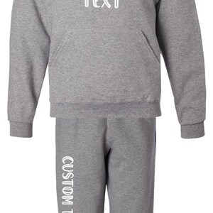 Pode incluir: Conjunto de moletom com capuz e cal&ccedil;as de moletom cinza. O moletom tem um bolso frontal e um capuz com cord&atilde;o. O texto "CUSTOM TEXT" &eacute; impresso em branco na frente do moletom e na perna esquerda das cal&ccedil;as. As cal&ccedil;as t&ecirc;m uma faixa azul marinho na lateral.