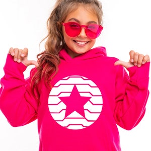 K&ouml;nnte beinhalten: Ein leuchtend pinkfarbener Kapuzenpullover mit einem wei&szlig;en Stern-Design in einem Kreis. Die Person im Bild tr&auml;gt eine rosa Sonnenbrille und zeigt auf den Hoodie. Ein auff&auml;lliges Kleidungsst&uuml;ck.