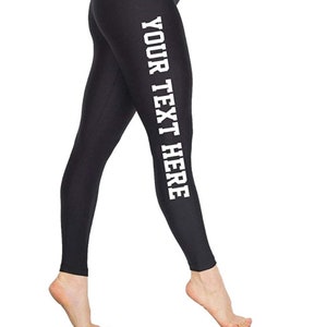 May include: Black leggings with the text "YOUR TEXT HERE" printed vertically in white on the left leg. The leggings are form-fitting and appear to be made of a smooth, opaque fabric. The person is standing on their toes.