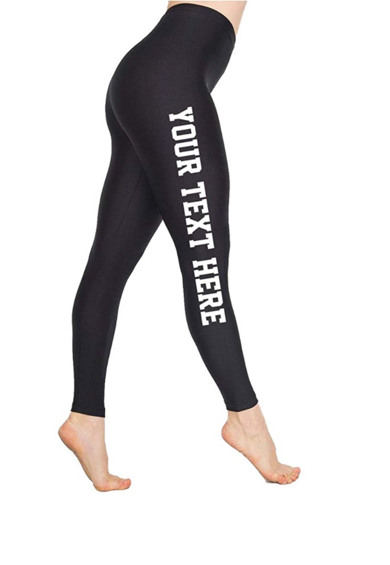 May include: Black leggings with the text "YOUR TEXT HERE" printed vertically in white on the left leg. The leggings are form-fitting and appear to be made of a smooth, opaque fabric. The person is standing on their toes.