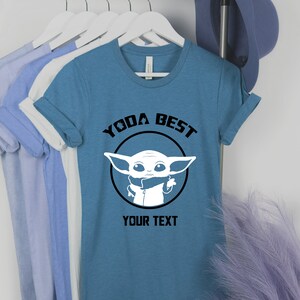 May include: A teal t-shirt with the text "YODA BEST" above a white graphic of a character from Star Wars. The text "YOUR TEXT" is below the graphic. The shirt is hanging on a rack with other shirts.