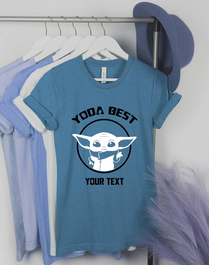 May include: A teal t-shirt with the text "YODA BEST" above a white graphic of a character from Star Wars. The text "YOUR TEXT" is below the graphic. The shirt is hanging on a rack with other shirts.