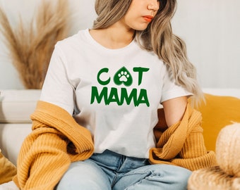 May include: White t-shirt with the green text "CAT MAMA". The letter "O" in "CAT" is replaced with a paw print inside a heart shape. The model is wearing blue jeans and a mustard-colored sweater.