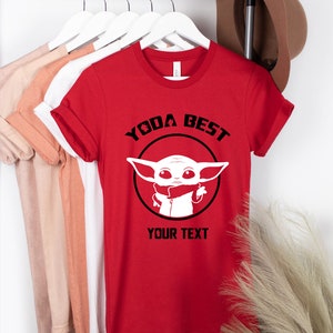 May include: Red t-shirt with the text "YODA BEST" above a white graphic of a character in a black circle. The text "YOUR TEXT" is below the graphic. Other t-shirts in various colors hang on a rack.