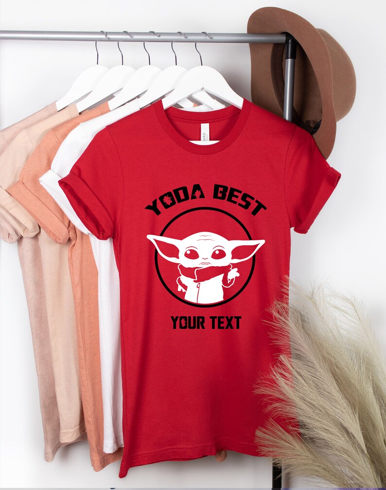 May include: Red t-shirt with the text "YODA BEST" above a white graphic of a character in a black circle. The text "YOUR TEXT" is below the graphic. Other t-shirts in various colors hang on a rack.