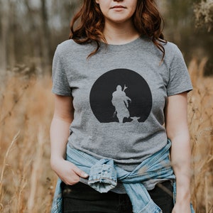May include: Gray t-shirt featuring a black silhouette design of a figure with a child-like figure in front of a black circle. The t-shirt is short-sleeved and made of a soft material. The person is wearing a denim jacket.