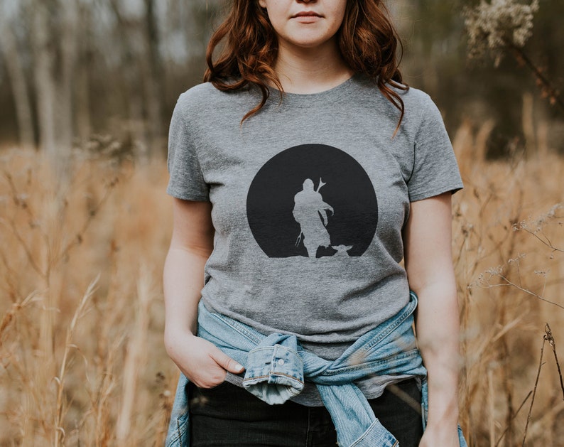 May include: Gray t-shirt featuring a black silhouette design of a figure with a child-like figure in front of a black circle. The t-shirt is short-sleeved and made of a soft material. The person is wearing a denim jacket.