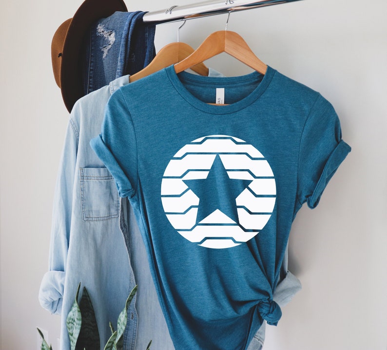 Puede incluir: Camiseta de color turquesa con un dise&ntilde;o de estrella blanca dentro de un c&iacute;rculo. La estrella es s&oacute;lida y el c&iacute;rculo est&aacute; formado por l&iacute;neas horizontales. La camiseta est&aacute; colgada en una percha de madera.