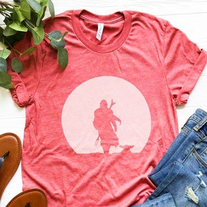 May include: Red t-shirt featuring a silhouette of a figure and a small creature against a light pink circle. The shirt is made of a soft material and is paired with blue jeans and brown sandals.