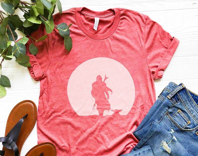 May include: Red t-shirt featuring a silhouette of a figure and a small creature against a light pink circle. The shirt is made of a soft material and is paired with blue jeans and brown sandals.