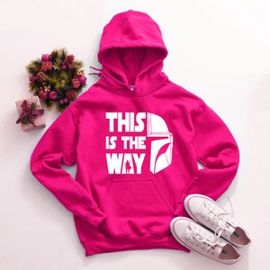 May include: A bright pink hooded sweatshirt with the text "THIS IS THE WAY" and a white helmet graphic. The sweatshirt has a front pocket and a drawstring hood. White sneakers are placed near the sweatshirt.