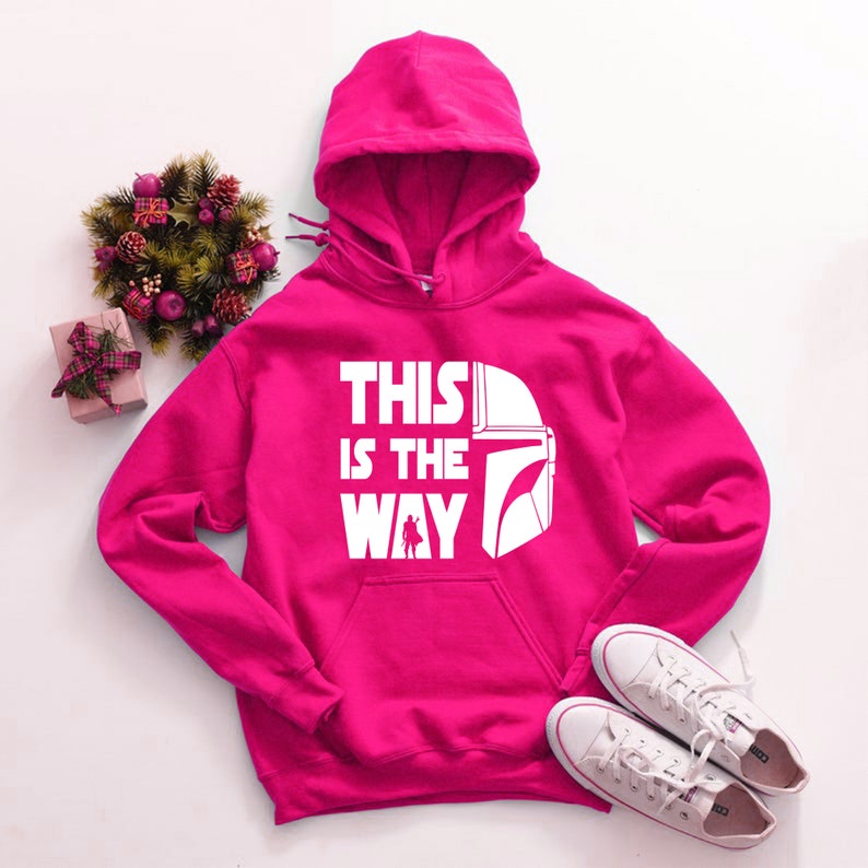 May include: A bright pink hooded sweatshirt with the text "THIS IS THE WAY" and a white helmet graphic. The sweatshirt has a front pocket and a drawstring hood. White sneakers are placed near the sweatshirt.