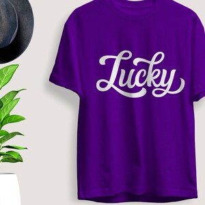 May include: A purple t-shirt with the word "Lucky" in white script. The shirt is displayed with a dark gray hat and a potted plant with green leaves.