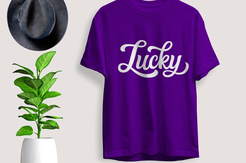 May include: A purple t-shirt with the word "Lucky" in white script. The shirt is displayed with a dark gray hat and a potted plant with green leaves.