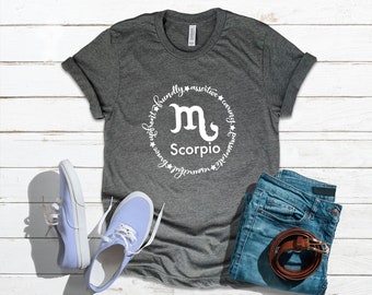 以下が含まれることがあります： 白い蠍座のシンボルと「Scorpio」の文字が円で囲まれた、濃い灰色のTシャツ。明るい青色のスニーカーと、茶色のベルトが付いた青色のジーンズも画像に写っています。