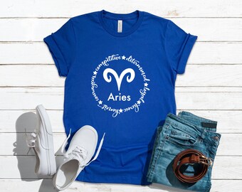 以下が含まれることがあります： 袖をまくったロイヤルブルーのTシャツ。白い牡羊座の星座シンボルと「競争力」や「決意」などの言葉が描かれています。青いジーンズ、茶色のベルト、白いスニーカーと組み合わせて、白い木の背景に配置されています。