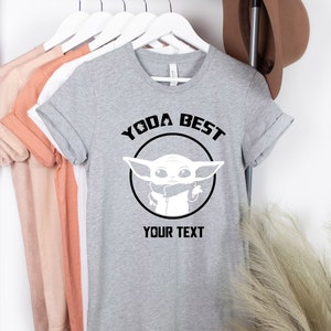 May include: A gray t-shirt with the text "YODA BEST" above a white graphic of a character from the Star Wars series. The text "YOUR TEXT" is below the graphic. The shirt is hanging on a rack with other shirts and a brown hat.