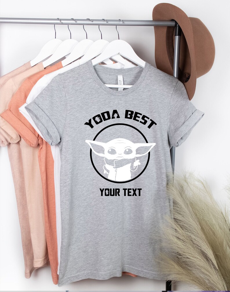 May include: A gray t-shirt with the text "YODA BEST" above a white graphic of a character from the Star Wars series. The text "YOUR TEXT" is below the graphic. The shirt is hanging on a rack with other shirts and a brown hat.