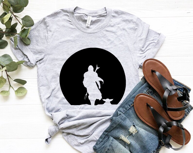 May include: A light gray t-shirt featuring a black silhouette of a character and a small creature against a black circle. The shirt is paired with blue jeans and brown sandals with black straps. The shirt is made of a soft material.