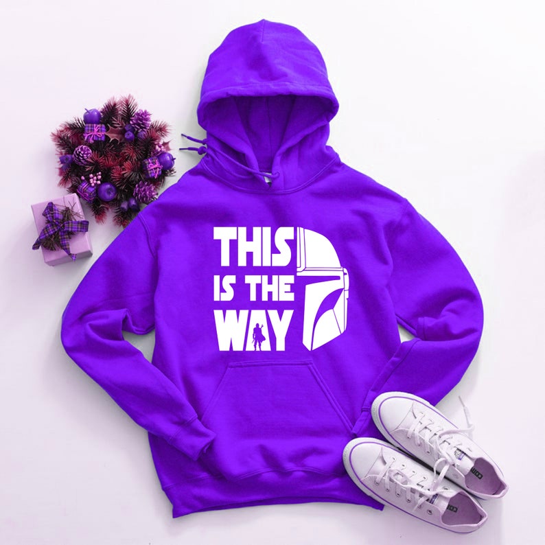 May include: A purple hooded sweatshirt with the text "THIS IS THE WAY" and a white graphic of a helmet and figure. White sneakers and a decorative floral arrangement are also in the image.
