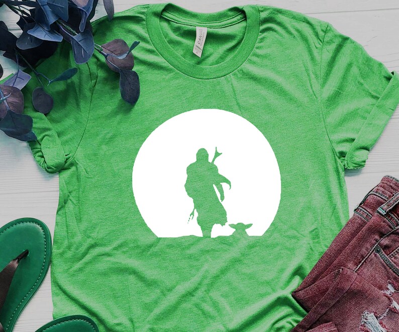 May include: Green t-shirt featuring a white silhouette design of a figure with a weapon, a small creature, and a large white circle in the background. The shirt is made of a soft material.