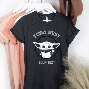 May include: Dark gray t-shirt with rolled sleeves. The shirt features a white graphic of a character with large ears inside a black circle, with the text "YODA BEST" above and "YOUR TEXT" below the graphic. Other shirts hang in the background.