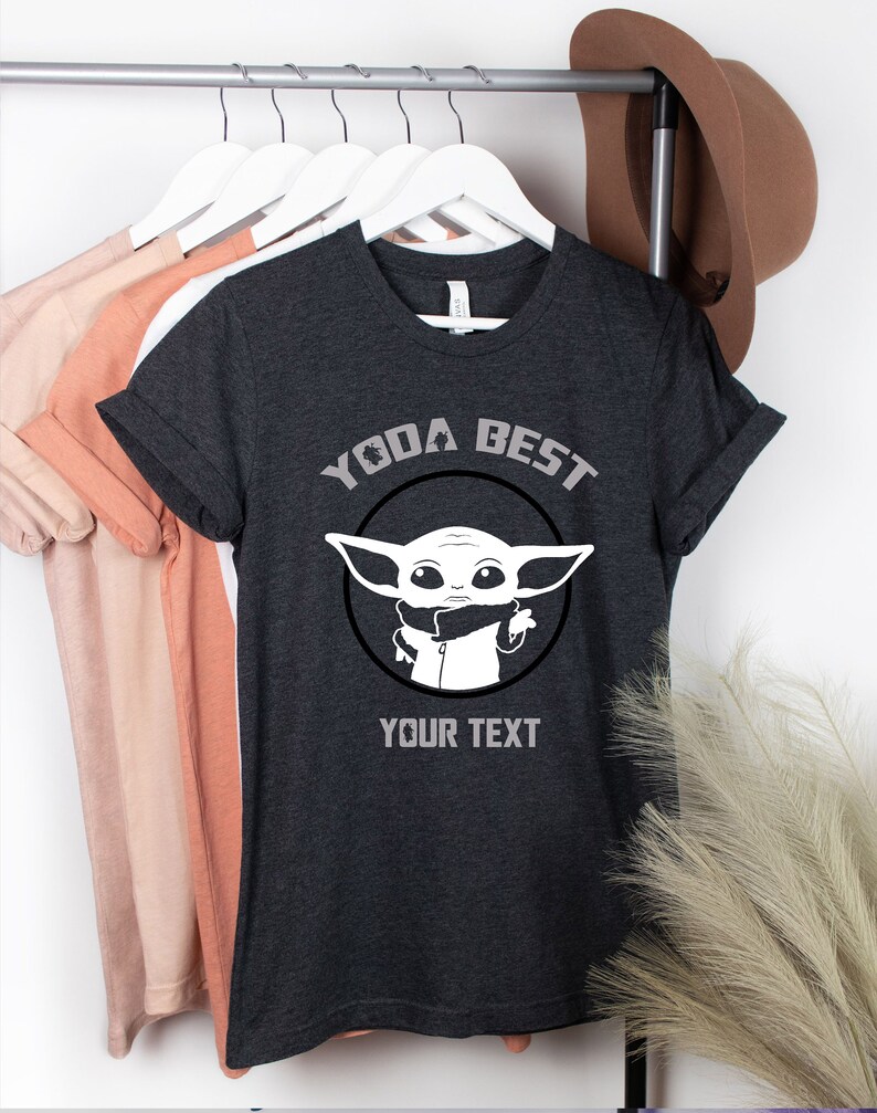 May include: Dark gray t-shirt with rolled sleeves. The shirt features a white graphic of a character with large ears inside a black circle, with the text "YODA BEST" above and "YOUR TEXT" below the graphic. Other shirts hang in the background.