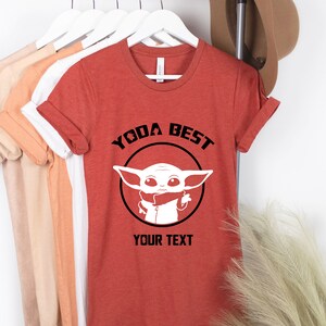 May include: Rust-colored t-shirt with the text "YODA BEST" above a white graphic of a character. Below the graphic is the text "YOUR TEXT". The shirt is hanging on a rack with other shirts.