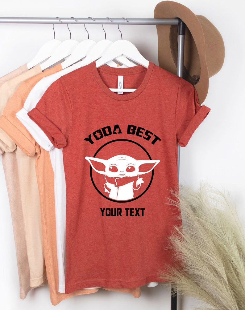 May include: Rust-colored t-shirt with the text "YODA BEST" above a white graphic of a character. Below the graphic is the text "YOUR TEXT". The shirt is hanging on a rack with other shirts.