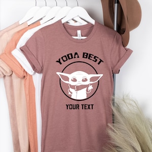 May include: Mauve t-shirt with rolled sleeves featuring a black and white graphic of a character with the text "YODA BEST" and "YOUR TEXT". Other t-shirts in various colors hang on a metal rack.