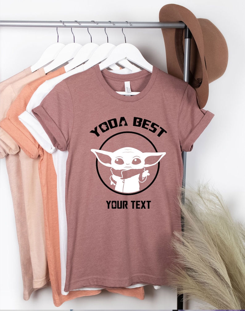 May include: Mauve t-shirt with rolled sleeves featuring a black and white graphic of a character with the text "YODA BEST" and "YOUR TEXT". Other t-shirts in various colors hang on a metal rack.