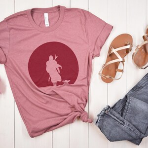 May include: A dusty rose t-shirt featuring a maroon graphic of a figure in front of a red circle. The shirt is paired with gold sandals and gray jeans. The shirt is made of a soft material.