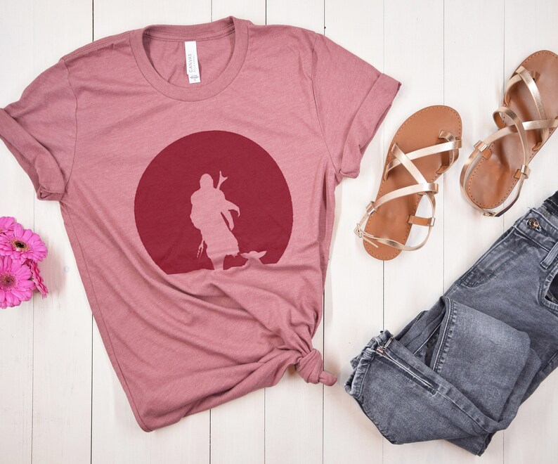 May include: A dusty rose t-shirt featuring a maroon graphic of a figure in front of a red circle. The shirt is paired with gold sandals and gray jeans. The shirt is made of a soft material.