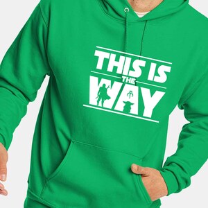 Puede incluir: Sudadera con capucha verde brillante con un gr&aacute;fico blanco que dice "THIS IS THE WAY". El dise&ntilde;o incluye siluetas de un personaje con capa, una criatura peque&ntilde;a y una figura con casco. Con bolsillo delantero.