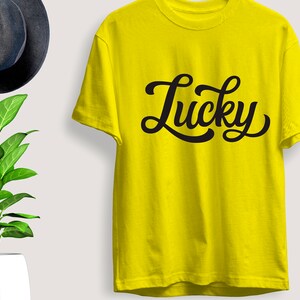 May include: A bright yellow t-shirt with the word "Lucky" in black cursive script. The shirt is short-sleeved and appears to be made of cotton. A dark gray fedora hat and a potted plant are in the background.