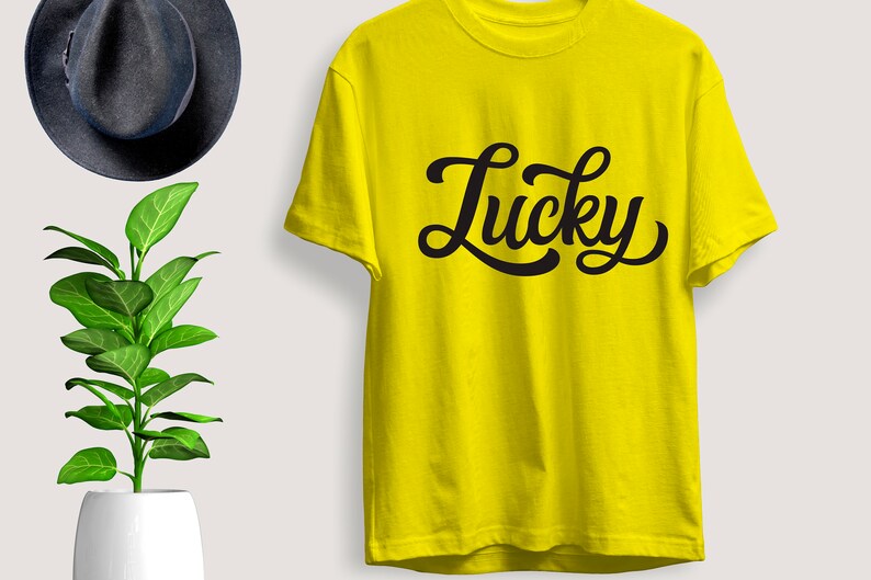 May include: A bright yellow t-shirt with the word "Lucky" in black cursive script. The shirt is short-sleeved and appears to be made of cotton. A dark gray fedora hat and a potted plant are in the background.