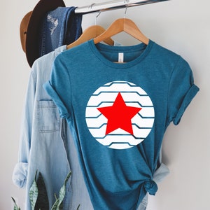 Puede incluir: Camiseta de color turquesa con un dise&ntilde;o de estrella roja dentro de un c&iacute;rculo a rayas blancas y azules. La camiseta se muestra en una percha de madera con otras prendas de vestir en el fondo. La camiseta es de un tejido suave.