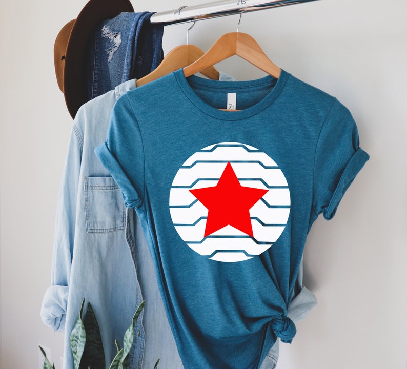 Puede incluir: Camiseta de color turquesa con un dise&ntilde;o de estrella roja dentro de un c&iacute;rculo a rayas blancas y azules. La camiseta se muestra en una percha de madera con otras prendas de vestir en el fondo. La camiseta es de un tejido suave.